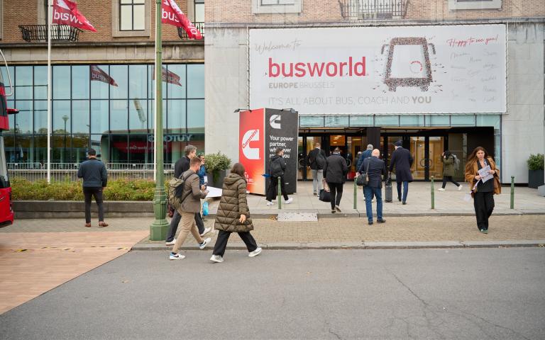 Visitors arriving at Busworld Europe 2025 in Brussels, Belgium, with Busworld flags and a welcome banner at the entrance to the international bus and coach exhibition.